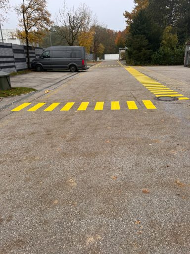 Linde, Oberschleißheim Gelber Zebrastreifen auf einem Parkplatz mit grauem Asphalt und parkenden Fahrzeugen.