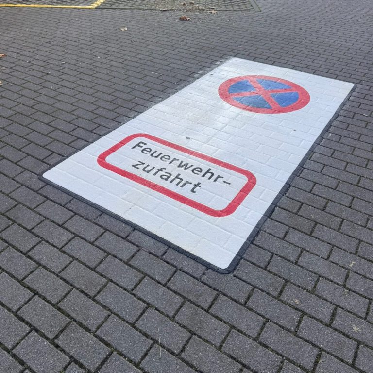 Feuerwehrzufahrten eine graue Pflasterfläche mit einem weißen Piktogramm, auf welchen "Feuerwehrzufahrt" steht und ein blau/rotes Parkverbotsschild