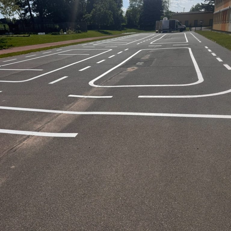 Weiß markierte Fahrbahnzeichen auf asphaltierter Fläche in einer Schulumgebung.